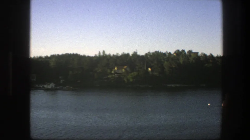 The moving boat perspective opens slightly onto rippled water, a dark green shoreline, and a few small structures partly hidden by trees.