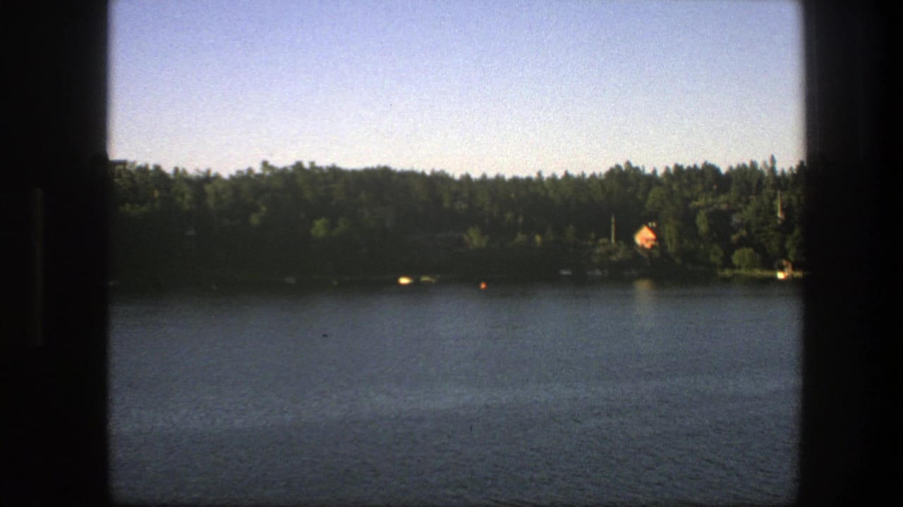 1975 Sweden 8mm lakeside view with calm water, dense trees, a distant orange-roofed house, pale sky, and dark frame borders.