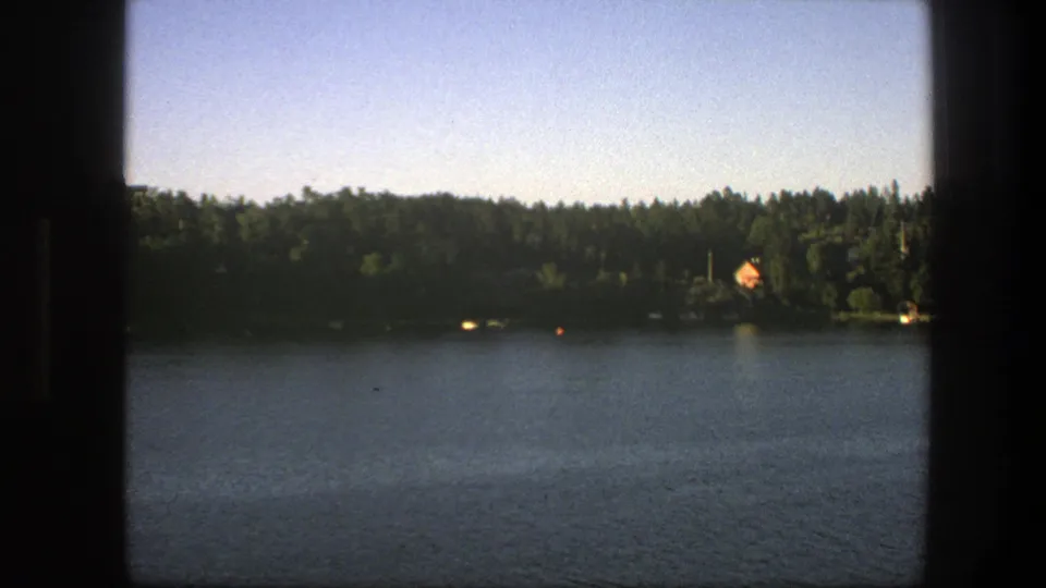 1975 Sweden 8mm lakeside view with calm water, dense trees, a distant orange-roofed house, pale sky, and dark frame borders.