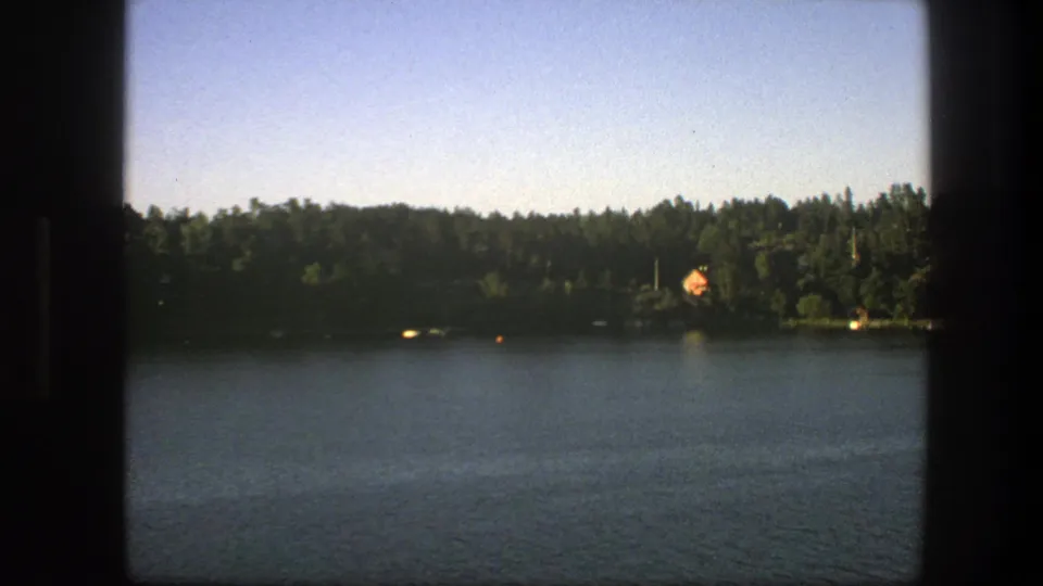 1975 Sweden home-movie frame showing lake water, wooded shoreline, small bright objects on the water, and a distant orange-roofed house.