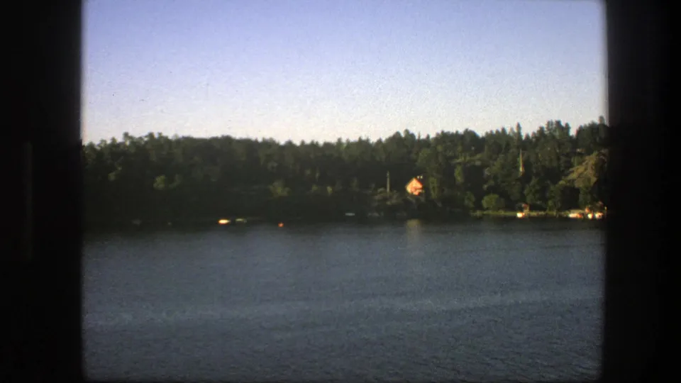 1975 Sweden 8mm view across a calm lake toward forest, a distant house, pale blue sky, and dark cabin or window edges.