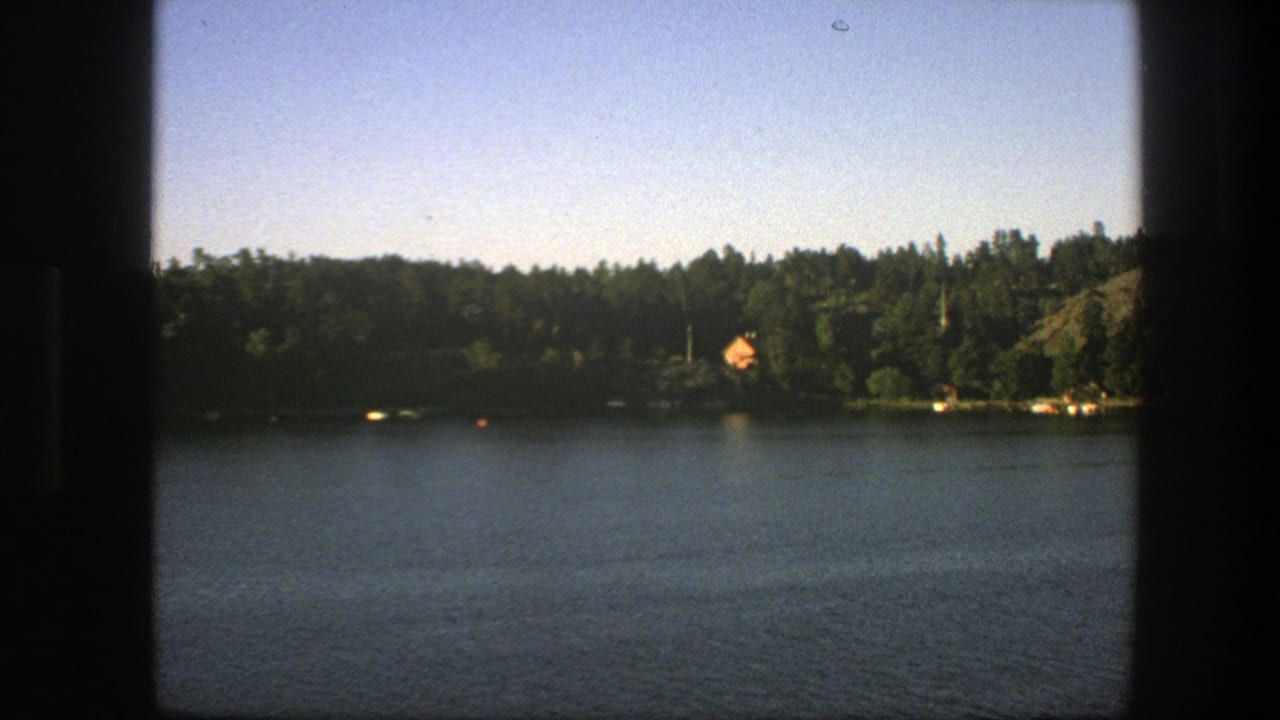Calm lake with forested shoreline, orange-roofed cabin, blue sky, and dark frame edges.
