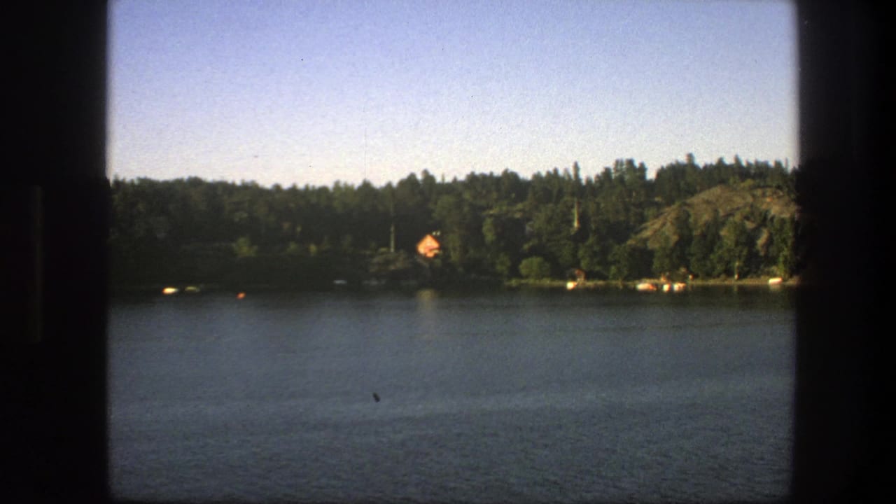 Wide lake view with wooded shoreline, small orange-roofed cabin, rocky bank, and dark vertical frame edge.
