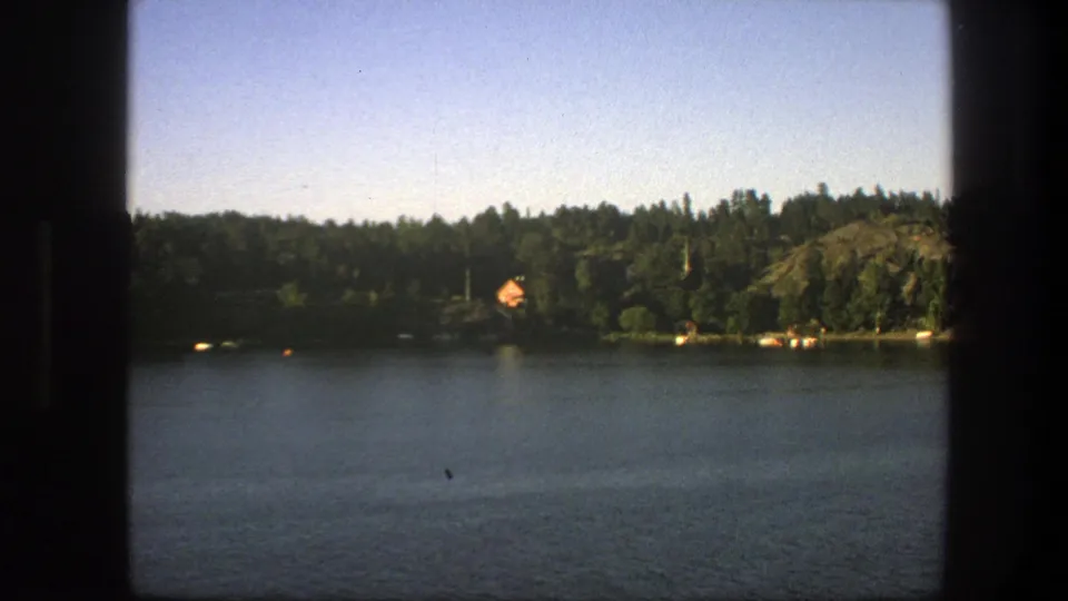 1975 Sweden lakeside home-movie frame showing still water, dense trees, a small cabin, and a dark central frame edge.