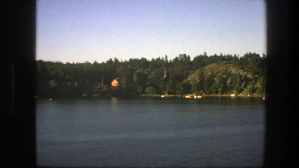 Swedish 1975 8mm frame with lake water in front of a wooded shore, orange-roofed cabin, rocky rise, and film gate edges.