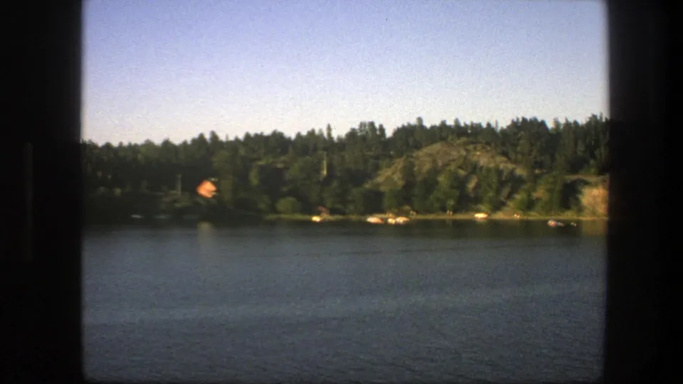 1975 Swedish lakeside frame showing calm water, tree-covered shore, rocky hillside, small shoreline objects, and dark frame edges.