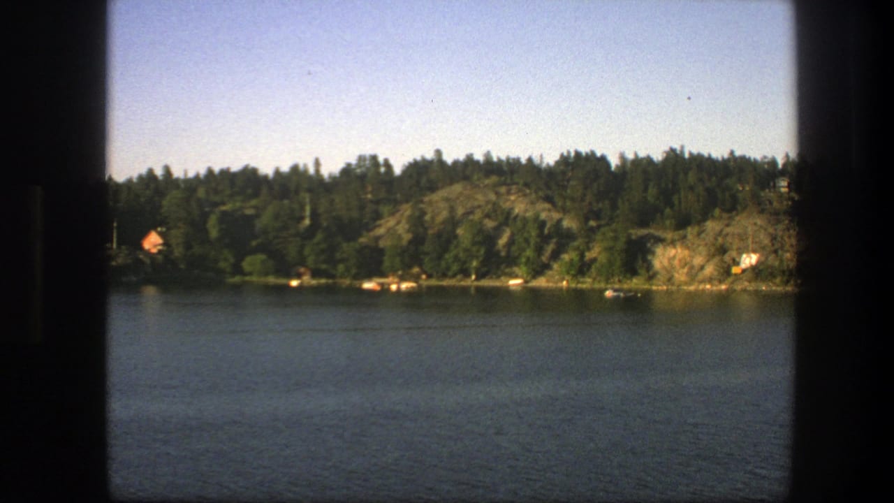 Forested lakeshore with rocky hills, distant houses, and water framed by dark vertical bars.