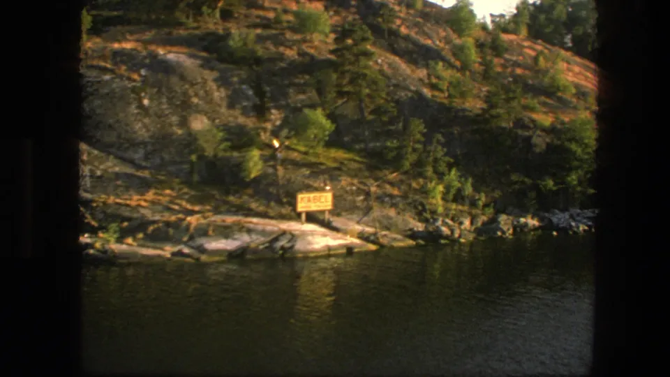 A closer 1975 Sweden 8mm view shows a rocky lakeside slope, green trees, and a yellow sign by the water.