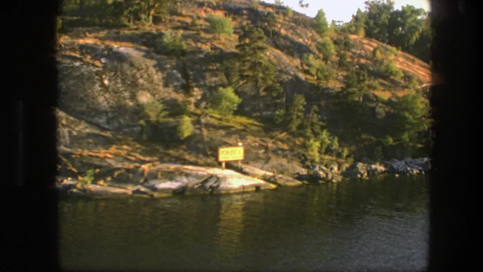 The final 1975 Sweden frame shows a yellow shoreline sign, green trees, and exposed rock beside calm lake water.