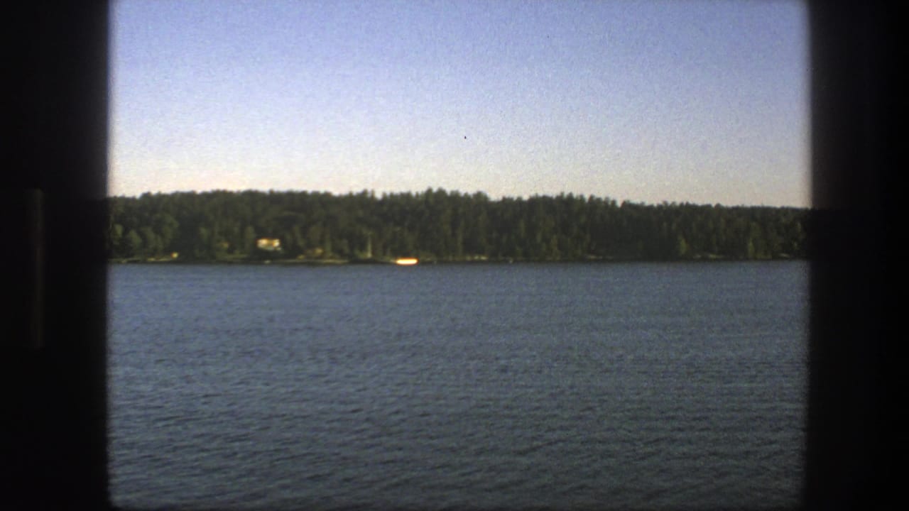 1975 Swedish 8mm frame looking across blue lake water toward a distant forested shore under a clear sky.