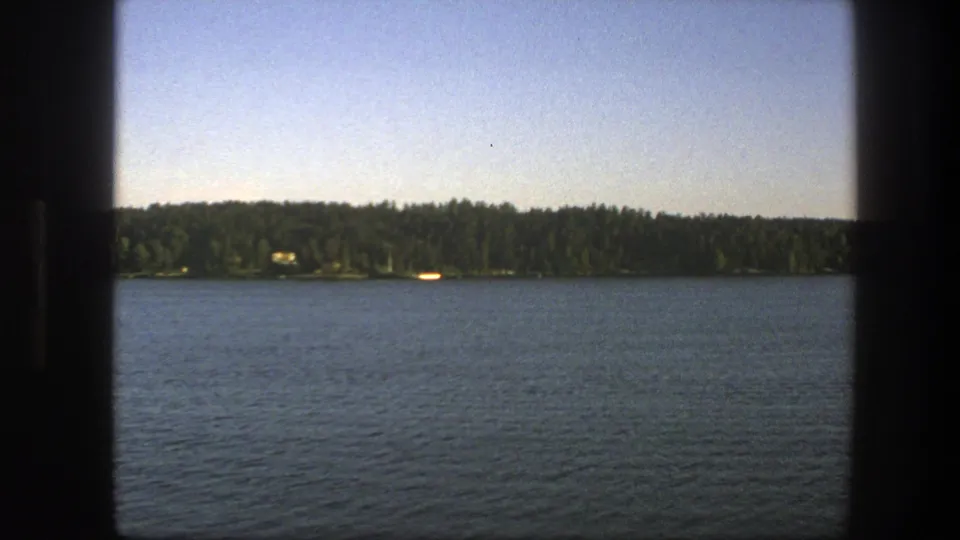 1975 Swedish 8mm frame looking across blue lake water toward a distant forested shore under a clear sky.