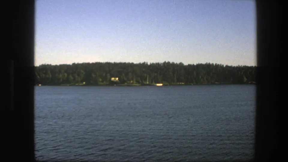 1975 Swedish 8mm frame showing calm blue water, a low forested horizon, pale sky, and small bright forms along the shore.
