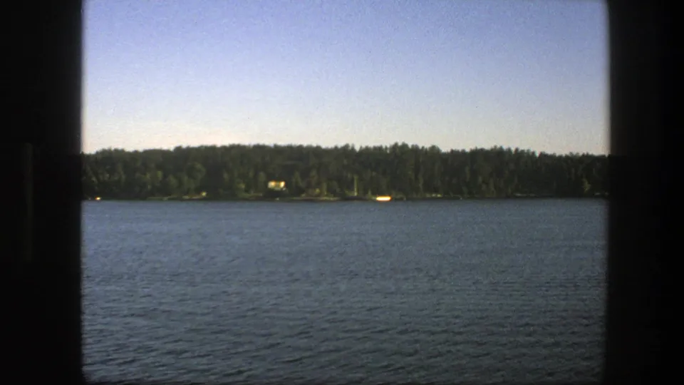 1975 Swedish 8mm frame showing blue lake water, dark window or railing edges, dense trees on the far shore, and a clear pale sky.