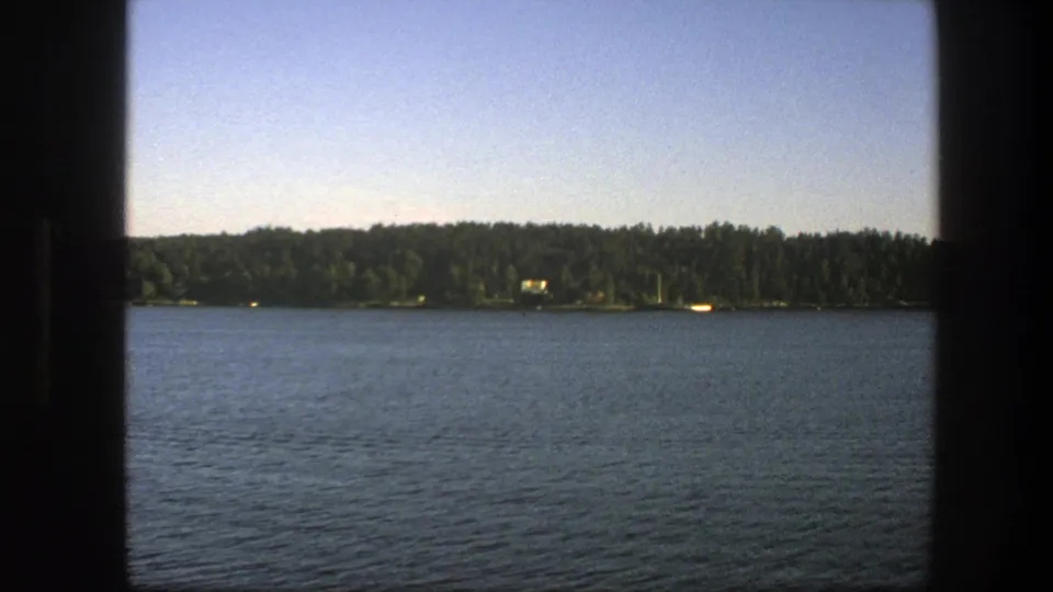 Swedish 1975 8mm still of a lake from a shaded framed viewpoint, with water in the foreground and a wooded shoreline beyond.
