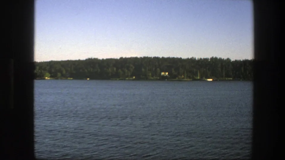 1975 Sweden 8mm frame showing a steady lakeside panorama with blue water, distant trees, small shore details, and clear sky.