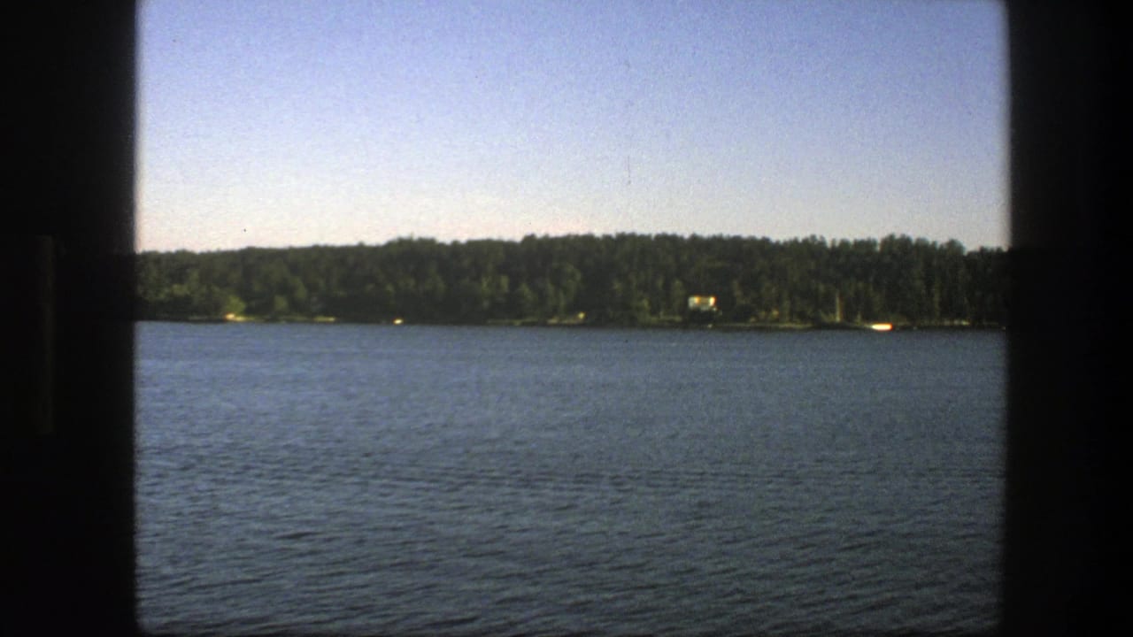 Calm blue lake with a distant forested shoreline under a clear sky, framed by dark film edges.