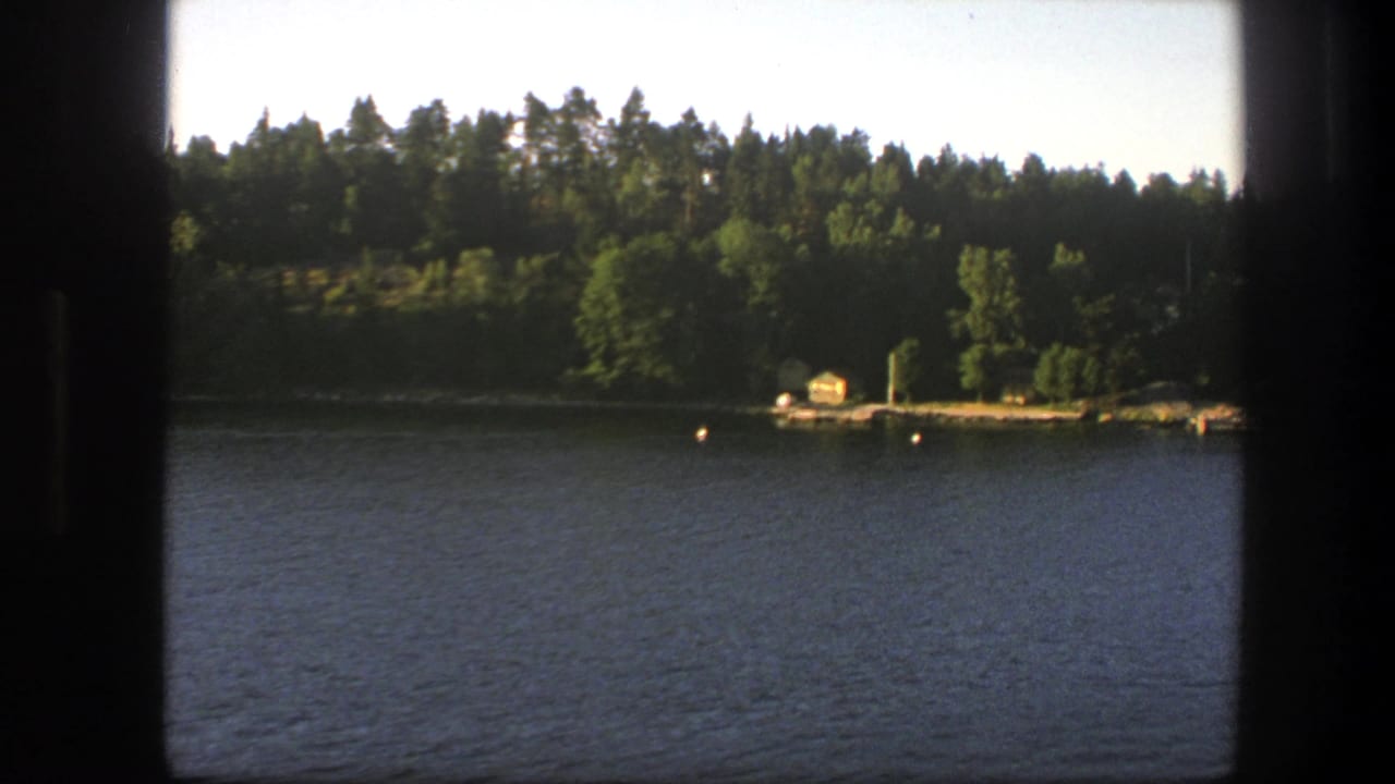 1975 Sweden lakeside view from a moving boat, showing trees, a small yellow boathouse, dock, and buoys across the water.