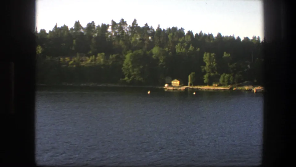 1975 Sweden lakeside view from a moving boat, showing trees, a small yellow boathouse, dock, and buoys across the water.