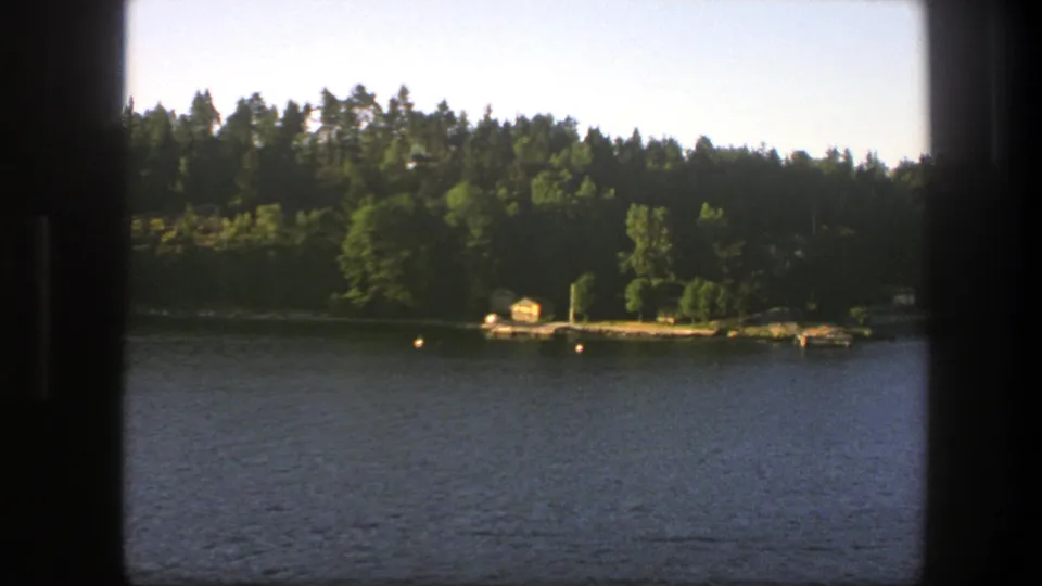 1975 Sweden home-movie frame of a wooded lake shore with calm water, buoys, dock, and a small yellow structure near the bank.