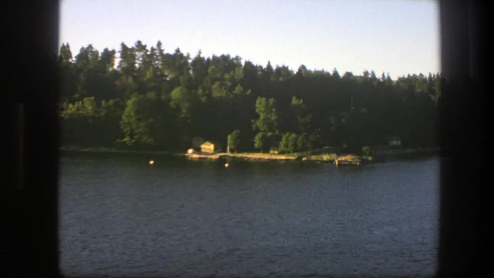 1975 Sweden 8mm boat-view frame with blue lake water, green forest, small buoys, a dock, and a yellow boathouse on shore.
