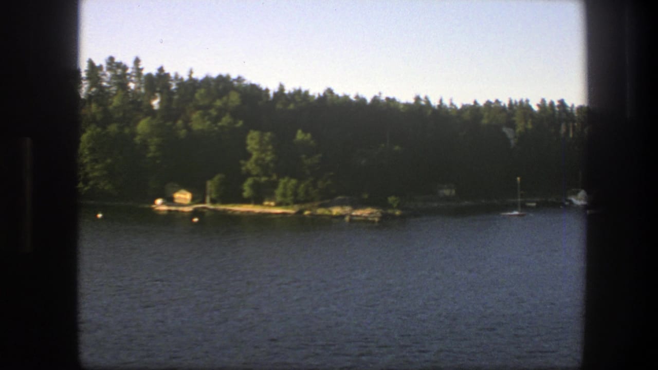 1975 Sweden home-movie frame showing lake water, forested shore, a small cabin, buoys, and a distant sailboat mast.