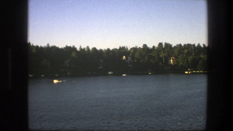 1975 Sweden 8mm lake view with a broad expanse of water, dark tree line, scattered cabins, and small boats near shore.