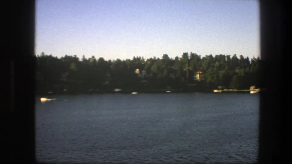 1975 Sweden home-movie frame of a lake crossing, with forested shore, small boats, and distant lakeside buildings in soft light.