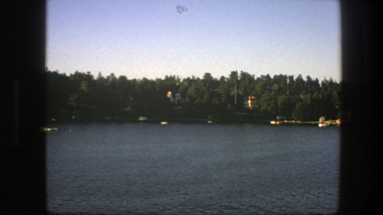 Boat-view across water toward a wooded Swedish shoreline with distant houses and docks.