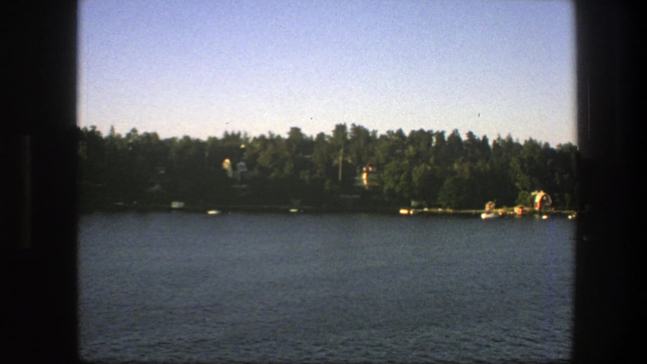 1975 Swedish home-movie frame showing calm water, wooded shoreline, distant houses, and small boats or docks from a boat.