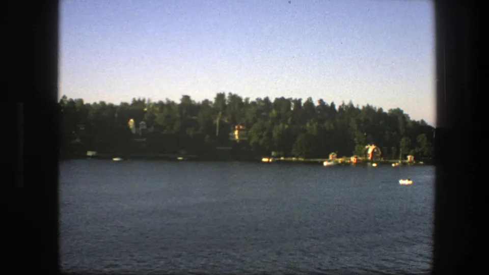 A 1975 8mm frame from Sweden shows a broad water surface, forested shore, dock area, and a small white boat in the distance.