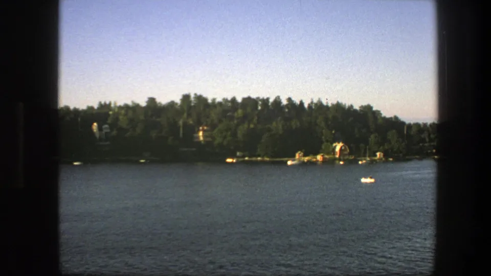Late-frame 1975 Swedish 8mm boat view with rippled water, wooded shoreline, small docks, distant houses, and a small white craft.