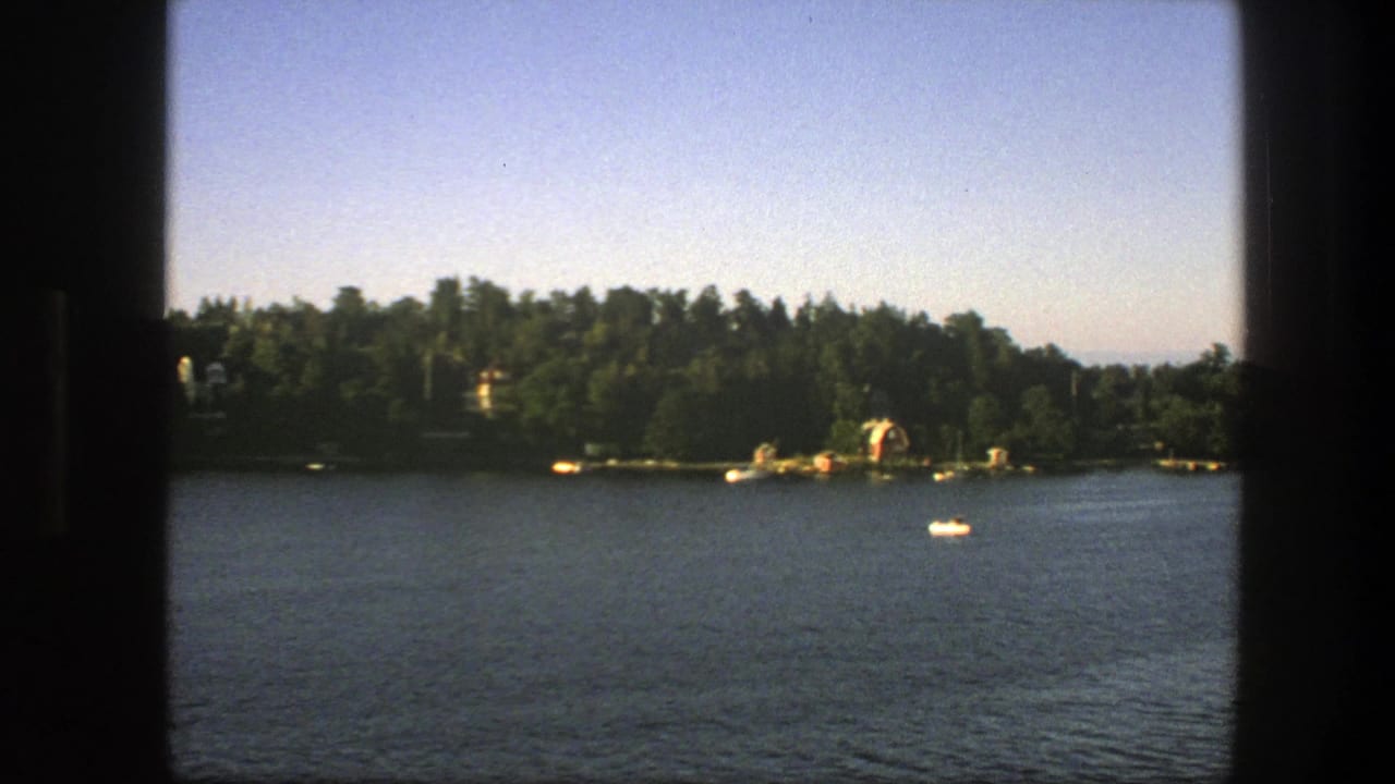 Small boat on a lake with wooded shoreline and lakeside buildings beyond.