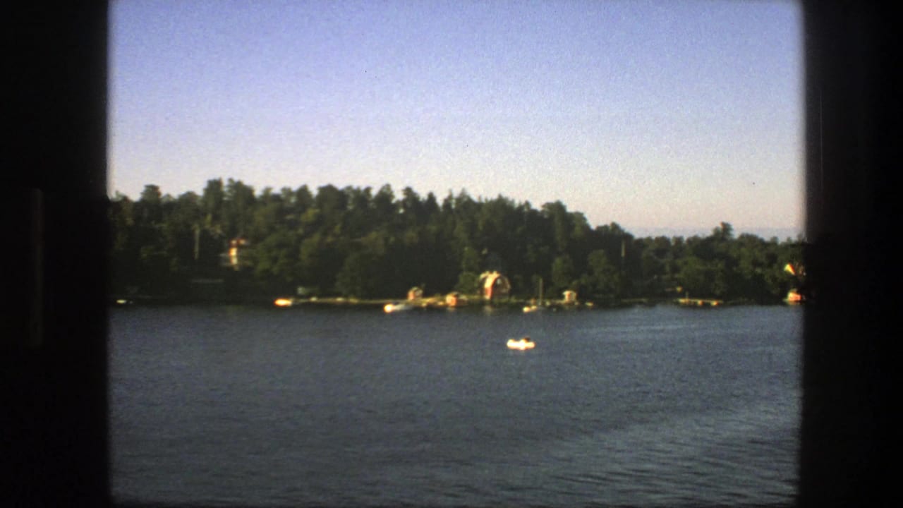 Small white boat sits on a Swedish lake in a 1975 home-movie frame, with dense trees and shoreline structures in the distance.