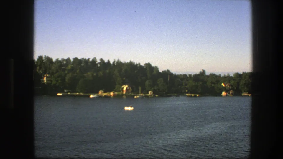 1975 Sweden lakeside frame with a single white boat on rippled water, framed by dark foreground edges and a wooded shore.