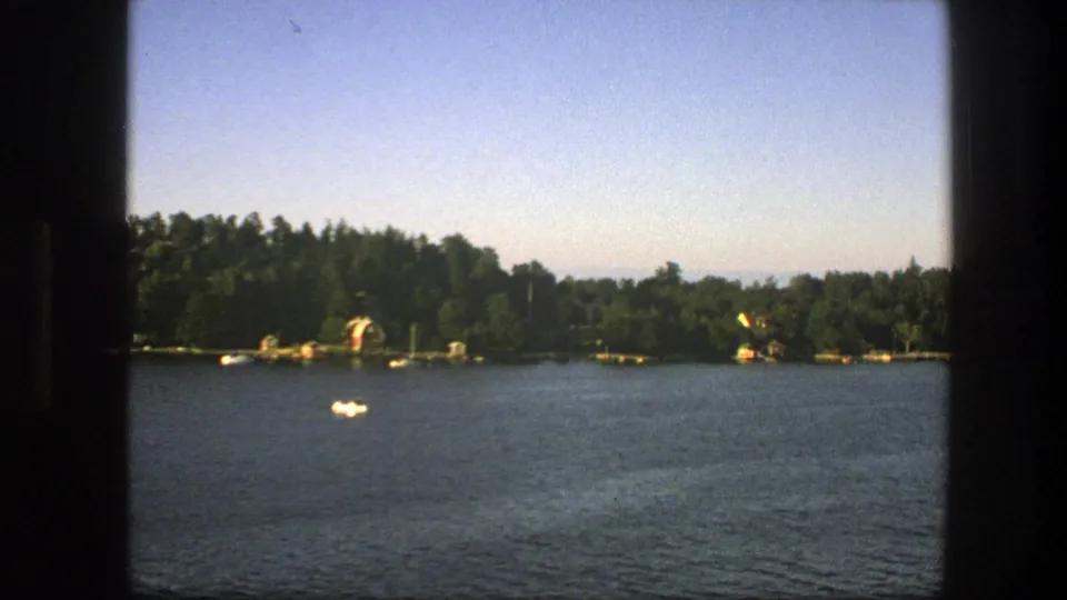 Final 1975 Swedish 8mm frame of a quiet lake, with a lone white boat, distant trees, scattered shoreline structures and soft film grain.