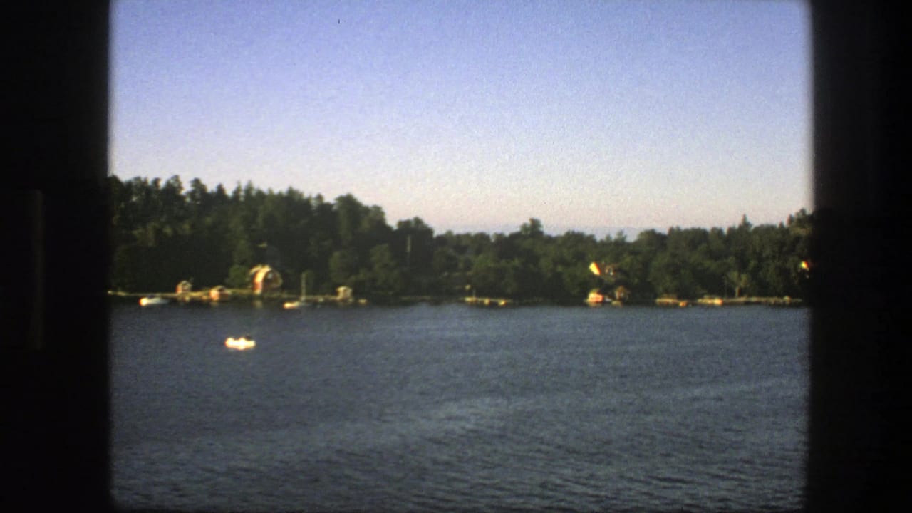 Lake or coastal water seen from a ferry, with a wooded shoreline and small boats in bright daylight.
