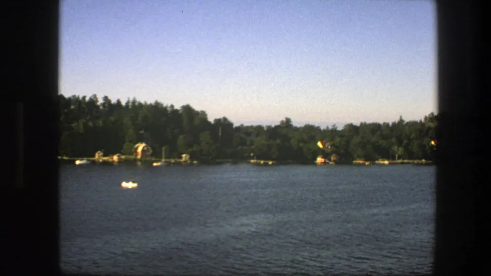 1975 Sweden 8mm view over calm water toward a tree-lined shore, with small boats visible under a pale blue sky.