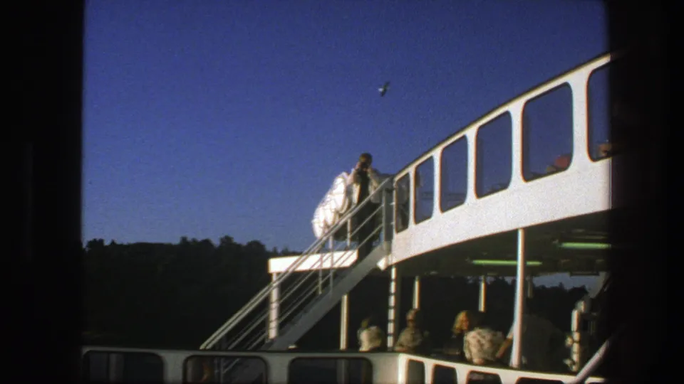 1975 Sweden 8mm frame of a man descending a ferry staircase while passengers sit on the lower deck in sunlight.