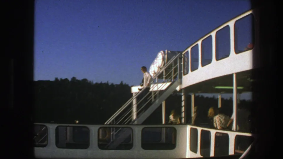 1975 Sweden ferry view with a man near the staircase, seated passengers below, and a wooded shoreline beyond.