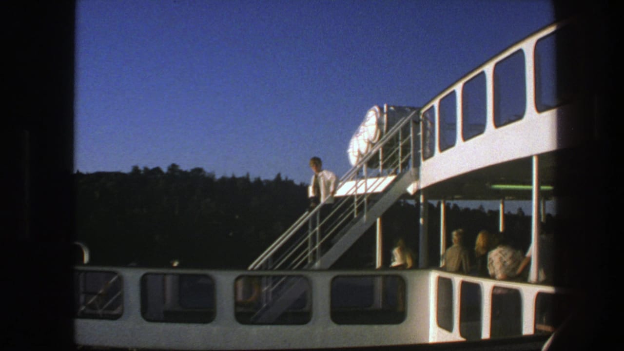 White ferry with exterior stairs, passengers on deck, and wooded shoreline behind it.