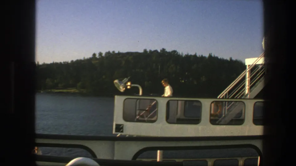 1975 Sweden ferry frame with a man in white on deck, exterior stairs, calm water, and forested shoreline.