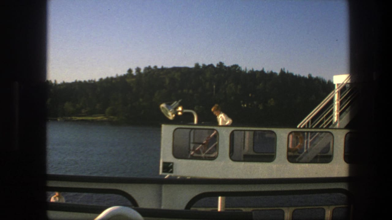 1975 Swedish 8mm frame showing a boat cabin with a person on deck beside calm lake water and a wooded shoreline.
