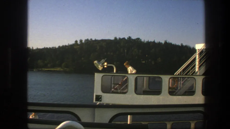 1975 Swedish 8mm frame showing a boat cabin with a person on deck beside calm lake water and a wooded shoreline.