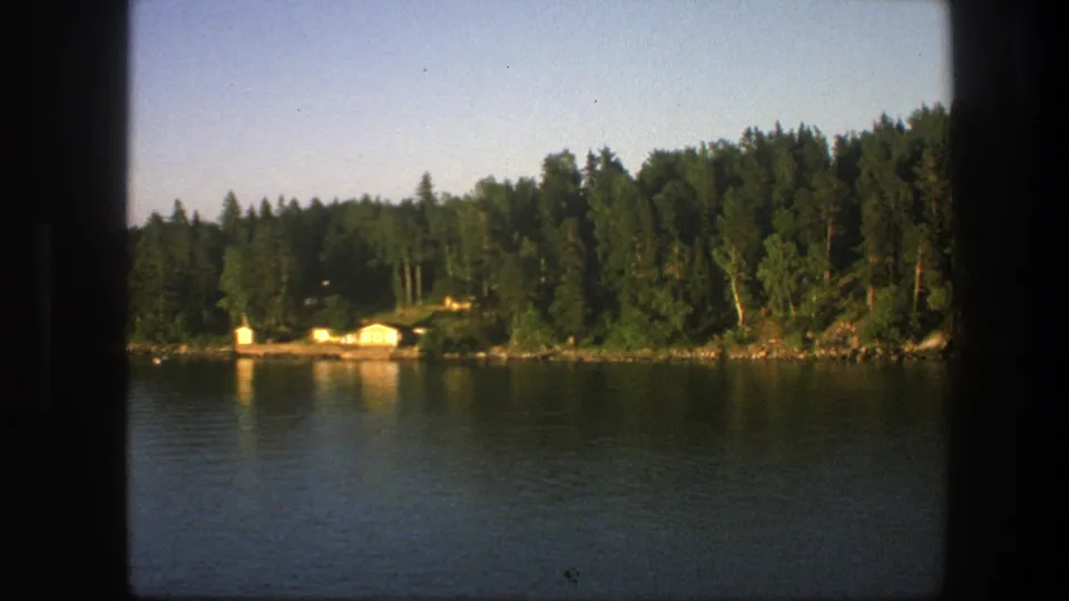 1975 Swedish lakeside view from a moving boat, with dense trees and small pale cabins along the water.