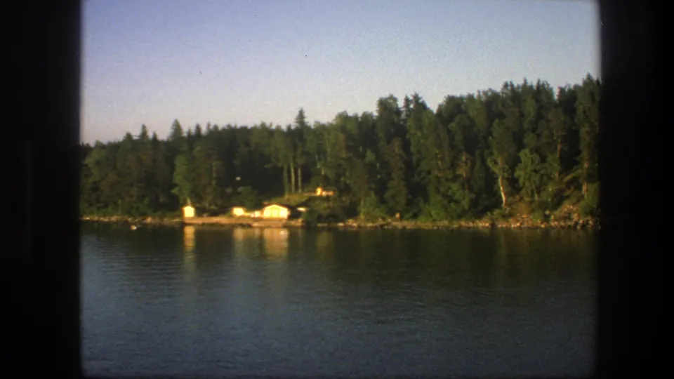 1975 Swedish 8mm frame of calm lake water, dense trees, and bright cabins on a wooded shoreline.