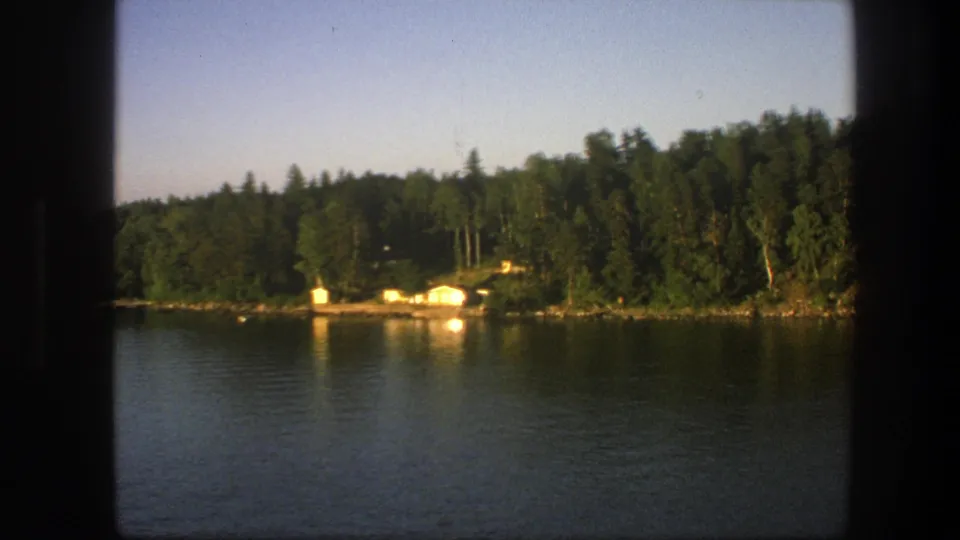 1975 Swedish home-movie frame showing a quiet lake crossing past forest, rocky shore, and small cabins.