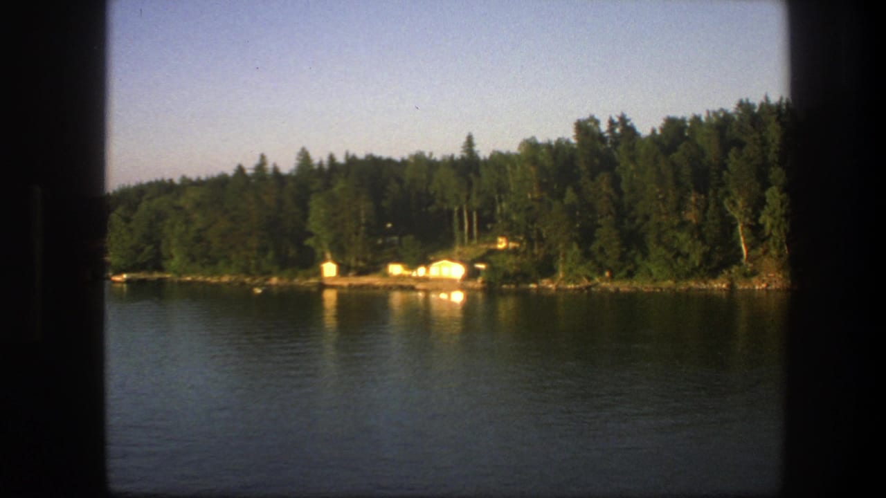 1975 Swedish 8mm frame showing calm lake water, forested shore, and bright cabins seen from a moving boat.