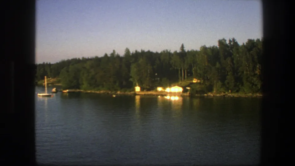A small sailboat sits near the Swedish forested shoreline, with cabins glowing in the background on 1975 8mm film.