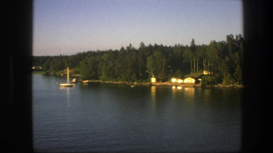 1975 Swedish lakeside view with a sailboat, reflective water, bright shoreline cabins, and dense green trees.