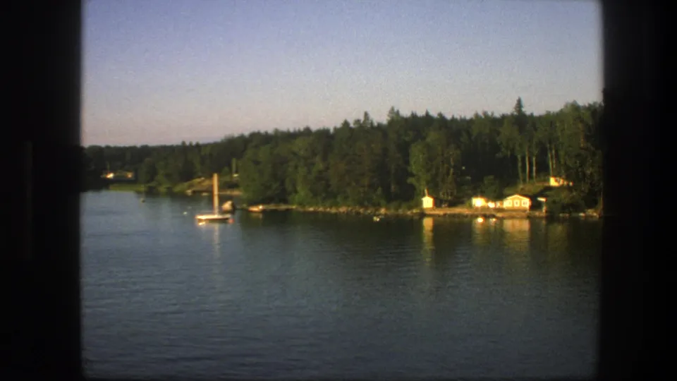 Wide 1975 8mm view of a Swedish lake, showing a sailboat on calm water and cabins tucked against a wooded shore.
